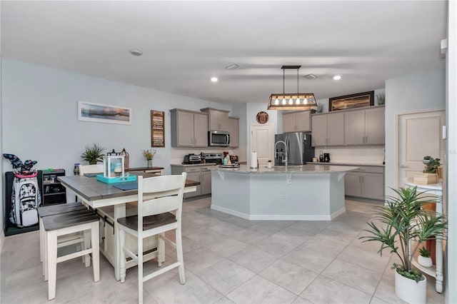 kitchen featuring gray cabinets, an island with sink, hanging light fixtures, light stone counters, and stainless steel appliances