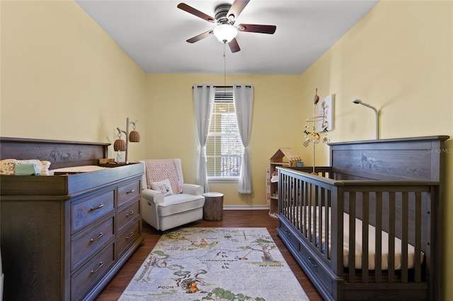 bedroom featuring a crib, a ceiling fan, baseboards, and dark wood-type flooring