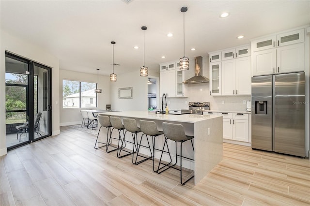 kitchen featuring pendant lighting, wall chimney range hood, appliances with stainless steel finishes, an island with sink, and white cabinets