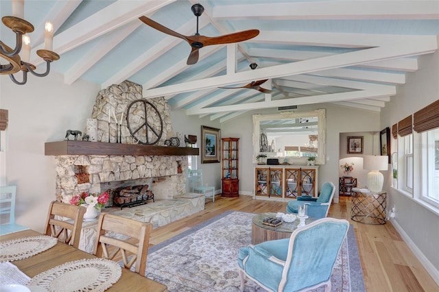 living room with ceiling fan with notable chandelier, a stone fireplace, lofted ceiling with beams, and wood finished floors