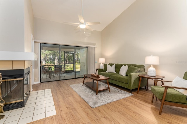 living room featuring ceiling fan, high vaulted ceiling, a tiled fireplace, and light wood-style floors