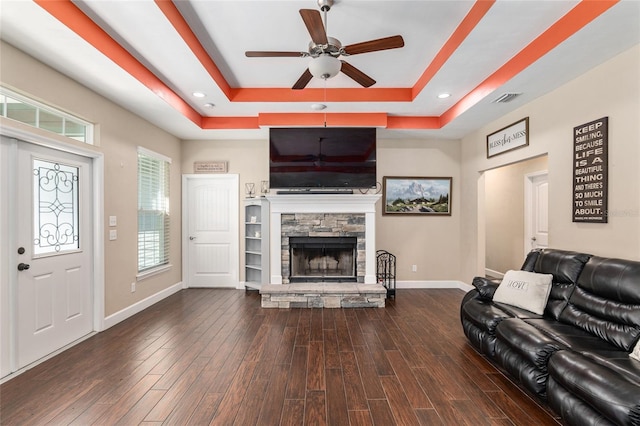 living room featuring a stone fireplace, dark wood-type flooring, visible vents, baseboards, and a tray ceiling