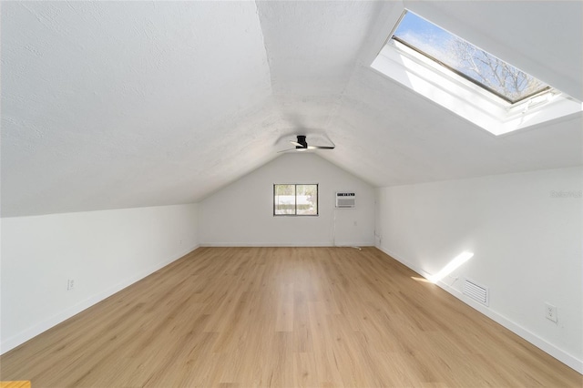 additional living space featuring vaulted ceiling with skylight, visible vents, a wall unit AC, a textured ceiling, and light wood-style floors