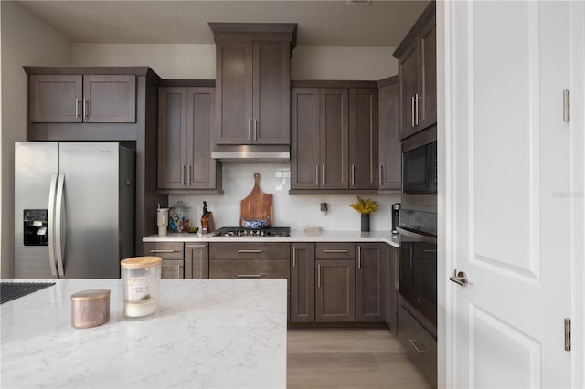 kitchen with dark brown cabinetry, decorative backsplash, appliances with stainless steel finishes, light stone counters, and under cabinet range hood