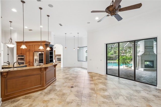 kitchen featuring pendant lighting, a ceiling fan, appliances with stainless steel finishes, brown cabinetry, and light stone countertops