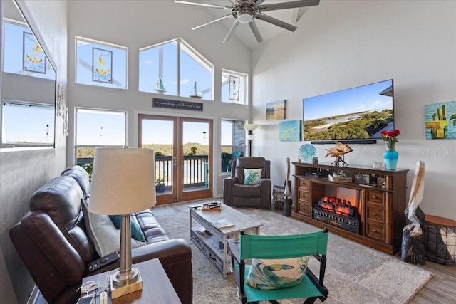 living room with french doors, a ceiling fan, wood finished floors, high vaulted ceiling, and a warm lit fireplace