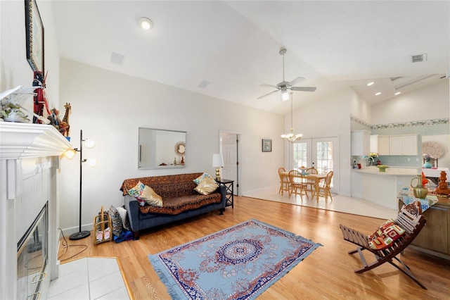 living room featuring light wood finished floors, visible vents, a tiled fireplace, high vaulted ceiling, and ceiling fan with notable chandelier