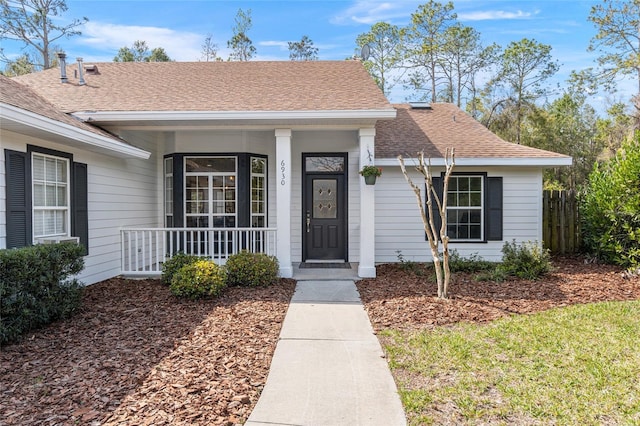 view of exterior entry featuring a shingled roof, covered porch, and fence