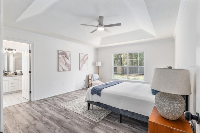 bedroom with crown molding, a raised ceiling, light wood-style floors, ensuite bath, and baseboards