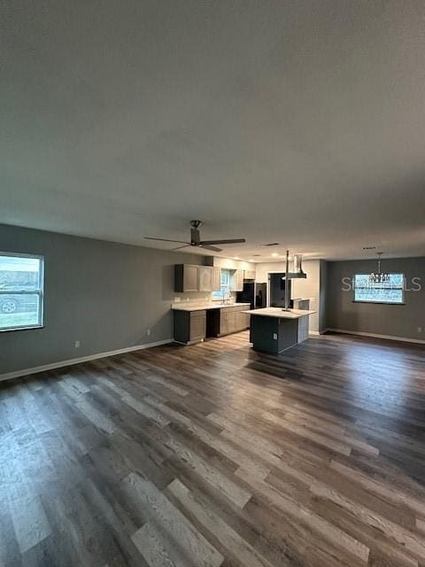 unfurnished living room featuring a wealth of natural light, dark wood-style flooring, baseboards, and ceiling fan with notable chandelier