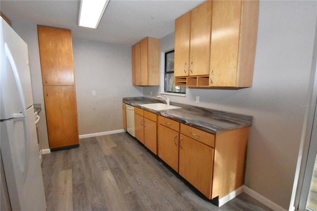 kitchen with dark countertops, a sink, wood finished floors, white appliances, and baseboards