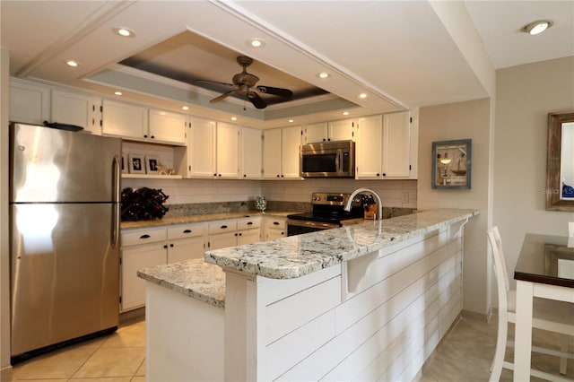 kitchen featuring a tray ceiling, appliances with stainless steel finishes, light tile patterned flooring, light stone countertops, and a peninsula