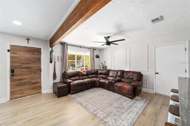 living room featuring light wood-style flooring, a decorative wall, visible vents, a ceiling fan, and beam ceiling