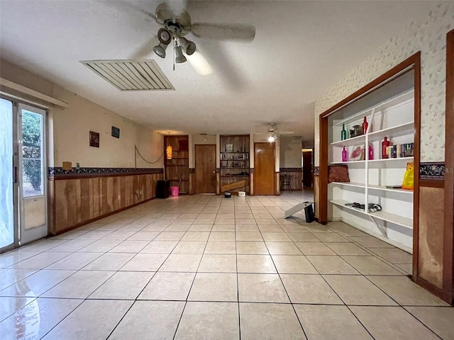 unfurnished room with light tile patterned floors, wooden walls, built in features, a ceiling fan, and a wainscoted wall