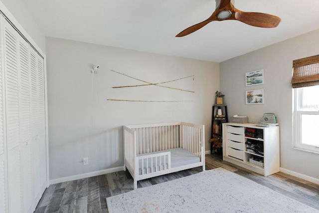 unfurnished bedroom featuring a crib, baseboards, wood finished floors, a closet, and a ceiling fan