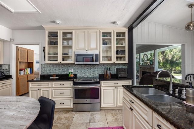 kitchen featuring stainless steel appliances, tasteful backsplash, cream cabinets, a sink, and dark stone counters