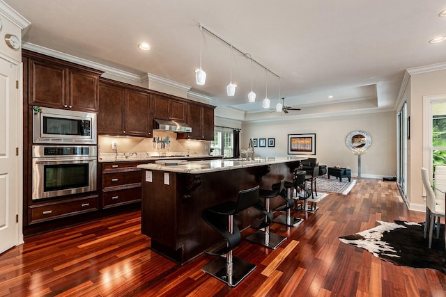 kitchen featuring under cabinet range hood, stainless steel appliances, dark wood-style flooring, a kitchen bar, and crown molding