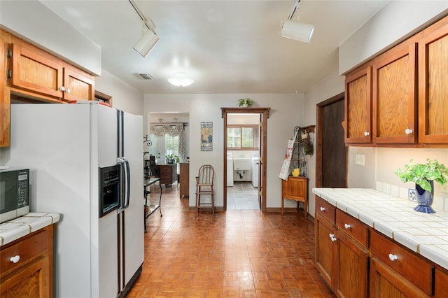 kitchen featuring tile counters, white refrigerator with ice dispenser, visible vents, and brown cabinetry