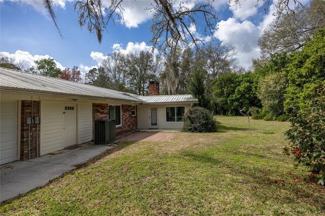 exterior space featuring brick siding, central AC unit, a lawn, a chimney, and metal roof