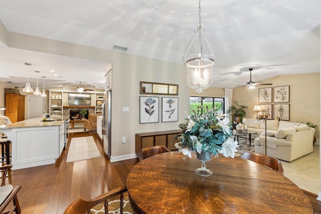 dining room featuring lofted ceiling, ceiling fan with notable chandelier, a fireplace, visible vents, and dark wood finished floors