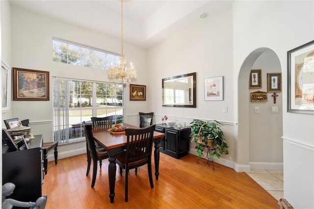 dining space featuring arched walkways, a notable chandelier, a towering ceiling, light wood-style floors, and baseboards