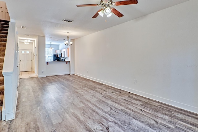 unfurnished living room featuring visible vents, ceiling fan with notable chandelier, stairway, light wood finished floors, and baseboards