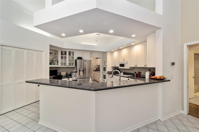 kitchen featuring white microwave, dark stone counters, a tray ceiling, stainless steel refrigerator with ice dispenser, and light tile patterned flooring