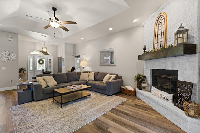 living area featuring a tray ceiling, a brick fireplace, wood finished floors, and a ceiling fan