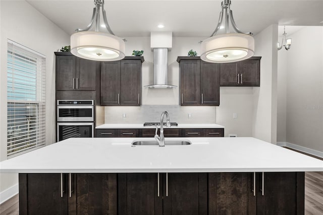 kitchen featuring light countertops, a sink, wood finished floors, stainless steel double oven, and wall chimney range hood