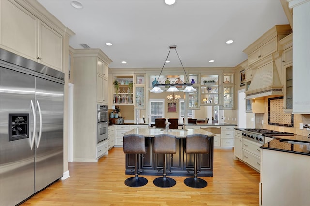 kitchen with a kitchen island with sink, custom range hood, light wood-style flooring, and appliances with stainless steel finishes