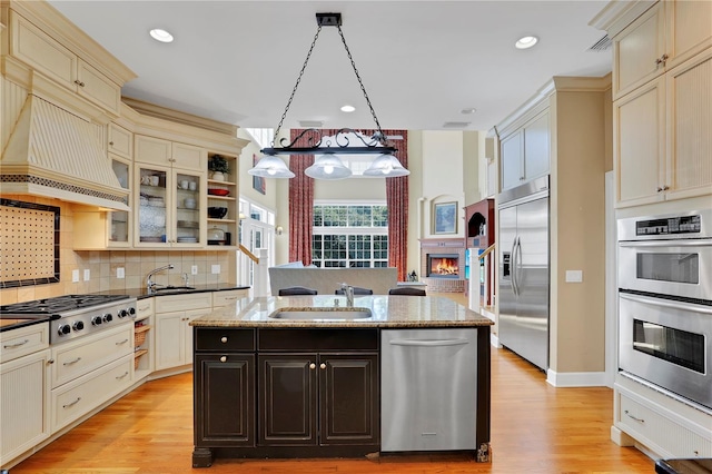 kitchen featuring appliances with stainless steel finishes, premium range hood, light wood-type flooring, a kitchen island with sink, and cream cabinetry