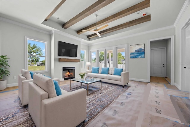 living room featuring baseboards, ornamental molding, a ceiling fan, beam ceiling, and a glass covered fireplace
