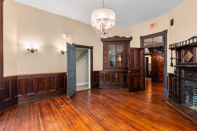 unfurnished dining area featuring a fireplace with flush hearth, dark wood-style floors, an inviting chandelier, and a wainscoted wall