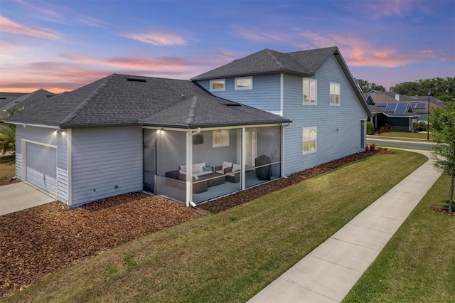back of property at dusk with roof with shingles, a lawn, a sunroom, concrete driveway, and an attached garage