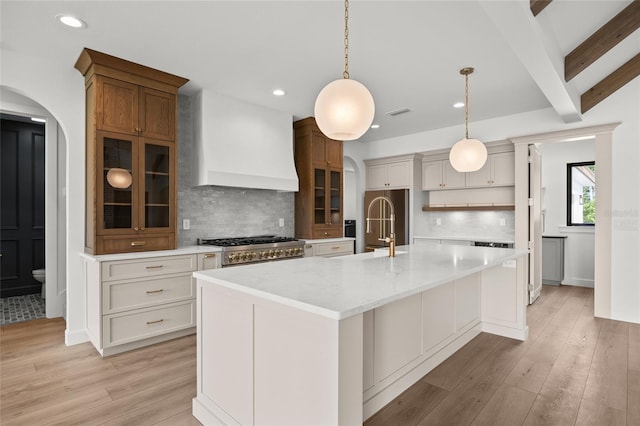 kitchen featuring light wood-style floors, arched walkways, a large island with sink, decorative backsplash, and custom range hood