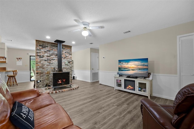 living area featuring a wood stove, visible vents, ceiling fan, wood finished floors, and wainscoting