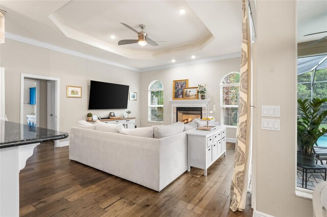 living area with a tray ceiling, a ceiling fan, dark wood-style flooring, and a warm lit fireplace