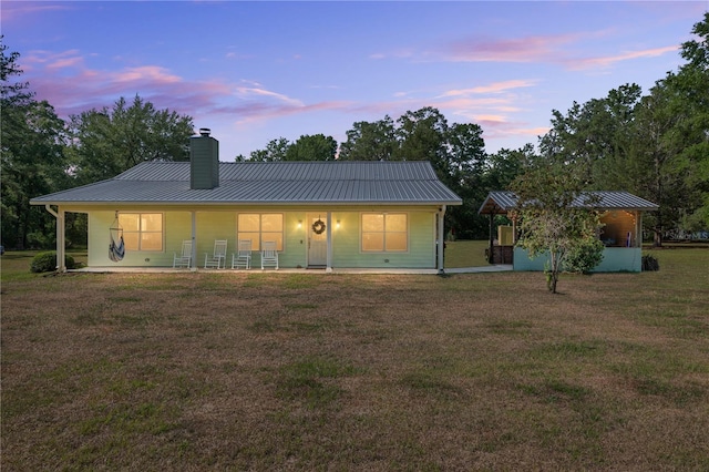 back of house at dusk with covered porch, a chimney, a standing seam roof, metal roof, and a lawn