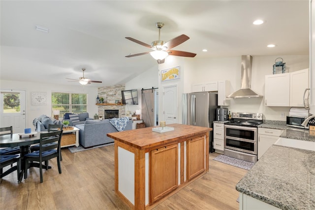 kitchen featuring ceiling fan, wall chimney exhaust hood, stainless steel appliances, light wood-type flooring, and a kitchen island