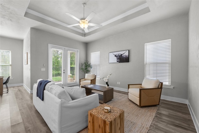 living area with a raised ceiling, wood finished floors, ceiling fan, baseboards, and crown molding