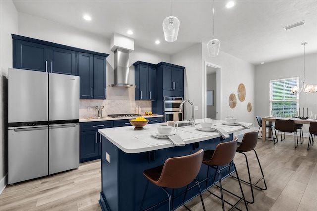 kitchen featuring stainless steel appliances, blue cabinetry, wall chimney range hood, light countertops, and an island with sink