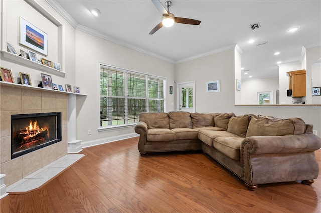 living room with ornamental molding, ceiling fan, wood finished floors, a tiled fireplace, and baseboards