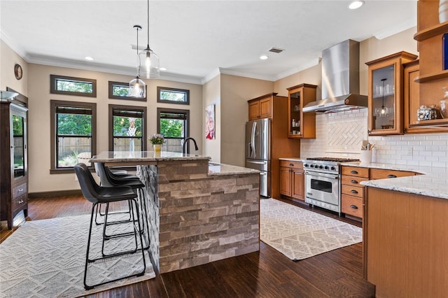 kitchen featuring wall chimney range hood, appliances with stainless steel finishes, brown cabinets, a sink, and light stone counters