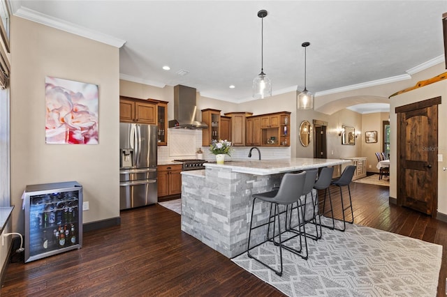 kitchen featuring stainless steel refrigerator with ice dispenser, wall chimney exhaust hood, brown cabinetry, arched walkways, and a kitchen bar