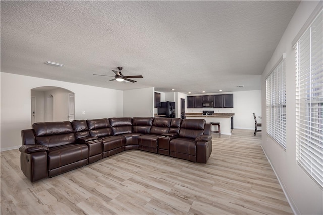 living room featuring arched walkways, light wood finished floors, a ceiling fan, baseboards, and a textured ceiling
