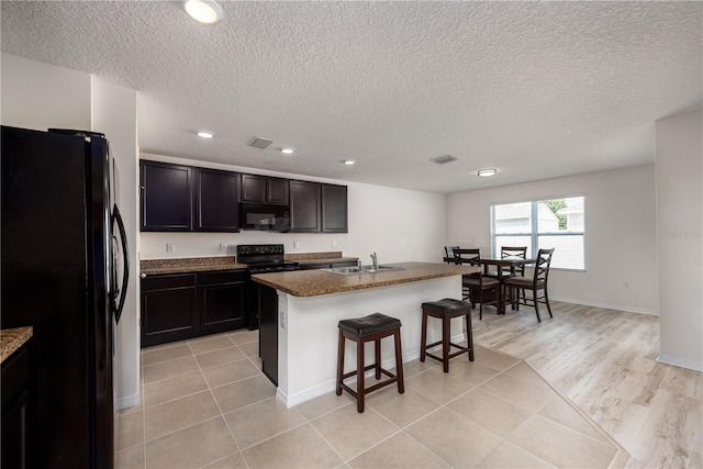 kitchen featuring black appliances, a sink, a kitchen bar, dark stone counters, and an island with sink