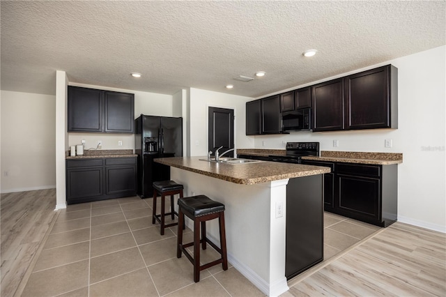 kitchen featuring black appliances, a sink, a center island with sink, a kitchen bar, and dark countertops
