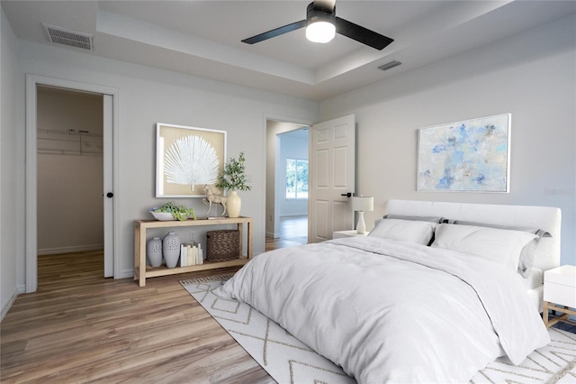 bedroom with a raised ceiling, a walk in closet, light wood finished floors, a closet, and a ceiling fan