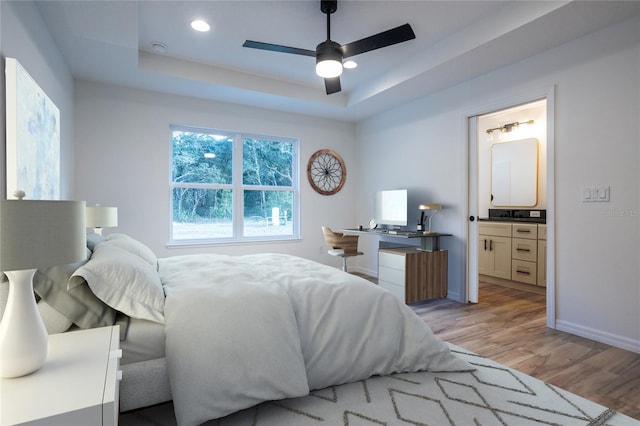 bedroom featuring light wood finished floors, a tray ceiling, baseboards, ensuite bathroom, and recessed lighting