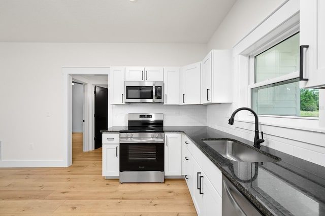 kitchen with appliances with stainless steel finishes, a sink, white cabinets, light wood-style floors, and dark stone countertops